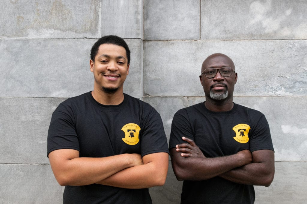 The two co-founders in their black Two Locals shirts with the yellow logo, standing with arms crossed.