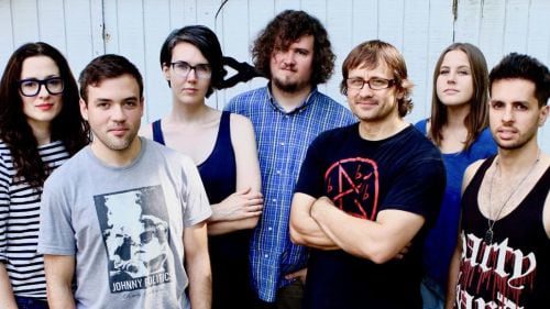 Wheatus - 7 band members stand, pictured waist-up, outside in front of a white wall.